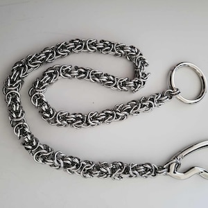K&ouml;nnte beinhalten: Eine silberfarbene Kette mit einem kreisf&ouml;rmigen Ring und einem gebogenen Verschluss. Die Kette besteht aus ineinandergreifenden Gliedern, wodurch ein strukturiertes Aussehen entsteht. Die Kette wird wahrscheinlich zum Sichern von Gegenst&auml;nden verwendet.