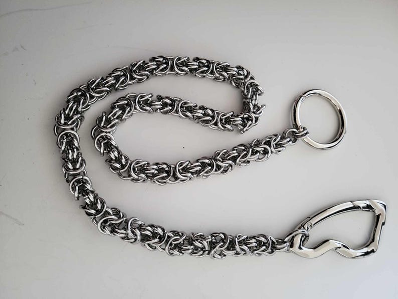 K&ouml;nnte beinhalten: Eine silberfarbene Kette mit einem kreisf&ouml;rmigen Ring und einem gebogenen Verschluss. Die Kette besteht aus ineinandergreifenden Gliedern, wodurch ein strukturiertes Aussehen entsteht. Die Kette wird wahrscheinlich zum Sichern von Gegenst&auml;nden verwendet.