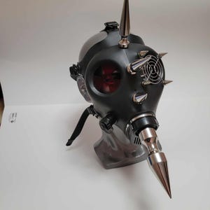 May include: A black gas mask with silver metal spikes and a long, pointed nose piece. The mask has a red-tinted lens and a small fan. The mask is displayed on a gray stand against a white background.