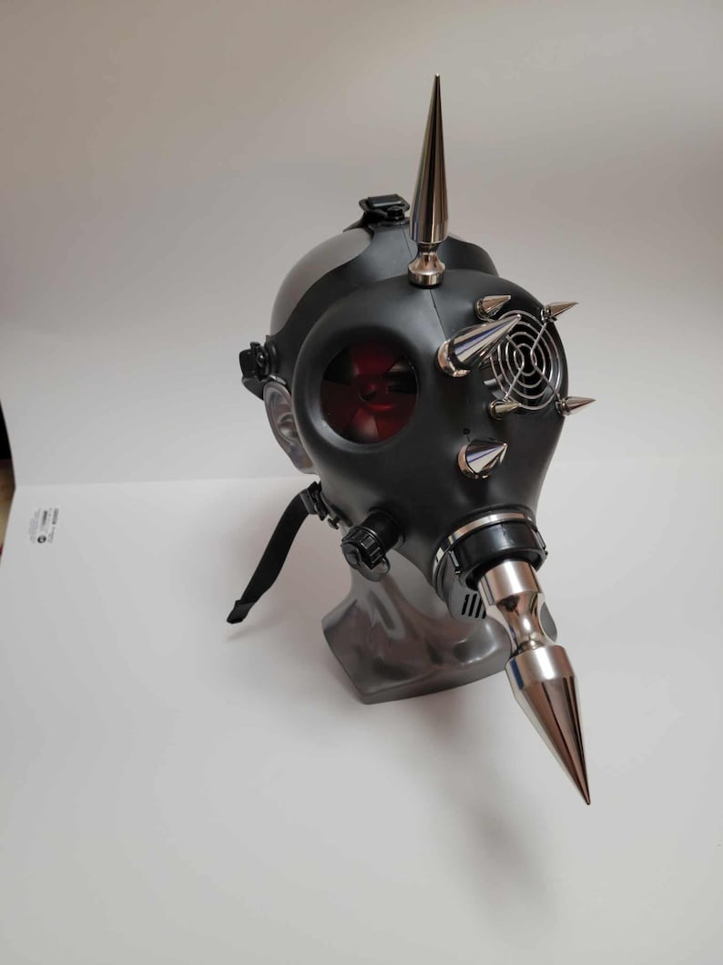 May include: A black gas mask with silver metal spikes and a long, pointed nose piece. The mask has a red-tinted lens and a small fan. The mask is displayed on a gray stand against a white background.