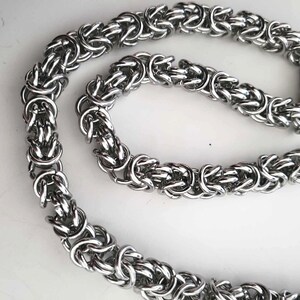 K&ouml;nnte beinhalten: Eine silberfarbene byzantinische Kettenhalskette. Die Kette besteht aus ineinandergreifenden Metallringen, die ein strukturiertes und detailliertes Muster bilden. Die Halskette wird auf einer wei&szlig;en Oberfl&auml;che pr&auml;sentiert.