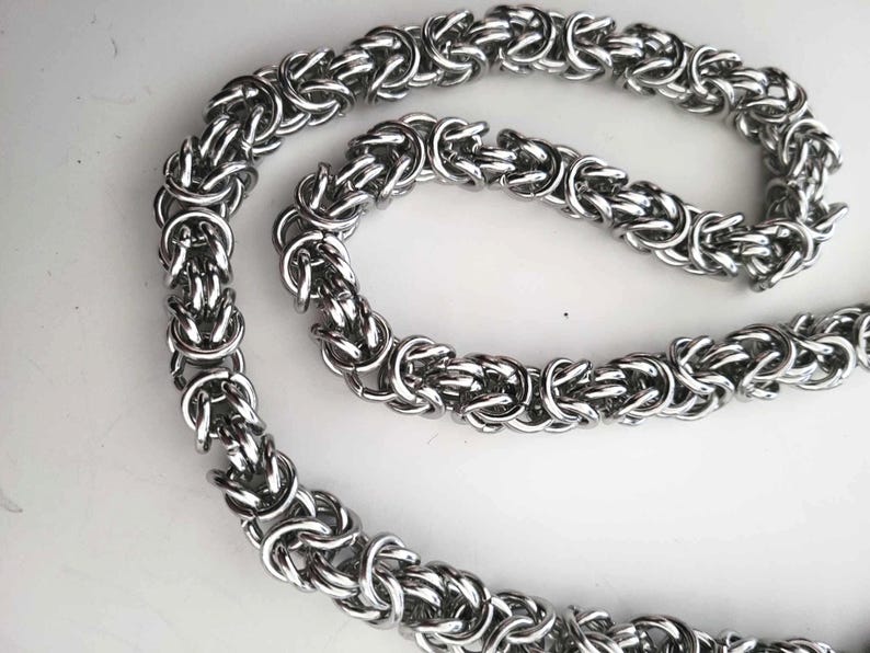 K&ouml;nnte beinhalten: Eine silberfarbene byzantinische Kettenhalskette. Die Kette besteht aus ineinandergreifenden Metallringen, die ein strukturiertes und detailliertes Muster bilden. Die Halskette wird auf einer wei&szlig;en Oberfl&auml;che pr&auml;sentiert.