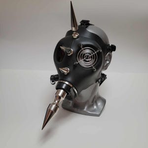 May include: A black gas mask with silver spikes and a silver nose cone. The mask has a silver fan on the front and is mounted on a gray mannequin head. The mask is a unique, industrial-style accessory.