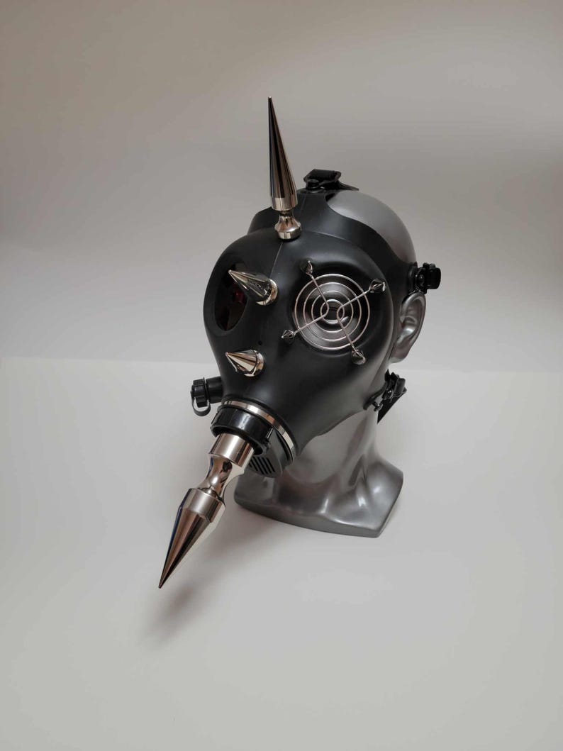 May include: A black gas mask with silver spikes and a silver nose cone. The mask has a silver fan on the front and is mounted on a gray mannequin head. The mask is a unique, industrial-style accessory.
