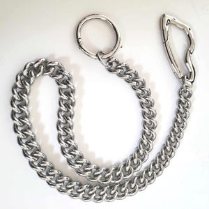 May include: A silver-colored chain with a large, circular ring at one end and a unique, curved clasp at the other. The chain links are thick and closely connected, creating a sturdy appearance. The item is displayed on a white background.