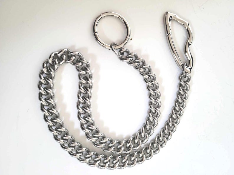 May include: A silver-colored chain with a large, circular ring at one end and a unique, curved clasp at the other. The chain links are thick and closely connected, creating a sturdy appearance. The item is displayed on a white background.