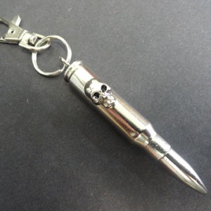 May include: Silver bullet keychain with a skull design. The bullet is attached to a silver key ring with a lobster clasp.