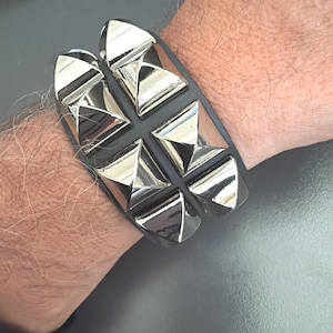 Puede incluir: Una pulsera negra con dos filas paralelas de tachuelas piramidales de metal plateado pulido. La pulsera tiene una base de material negro mate y liso, con un diseño estructurado que muestra cuatro acentos metálicos en forma de pirámide.