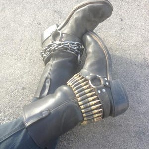 Puede incluir: Un par de botas de cuero negro con cadenas plateadas y un cinturón de balas. Las botas están gastadas y tienen un aspecto desgastado.