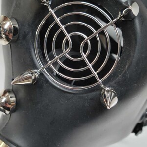 May include: Black gas mask with silver metal accents. The mask features a circular fan cover and multiple silver spikes. The mask has a strap for securing it to the face. The mask is made of a matte black material.