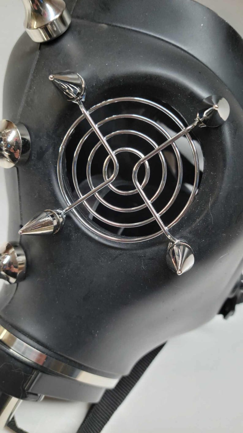 May include: Black gas mask with silver metal accents. The mask features a circular fan cover and multiple silver spikes. The mask has a strap for securing it to the face. The mask is made of a matte black material.