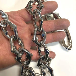 May include: A shiny silver chain with unique, stylized links. The links have a distinctive shape, and the chain is held up against a white background. The chain is made of a reflective metal.