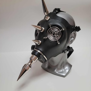 Peut inclure: Un masque à gaz noir avec des pointes et des détails en métal argenté. Le masque présente un design de ventilateur circulaire à l'avant et est présenté sur une tête de mannequin grise. Le masque a des sangles réglables.