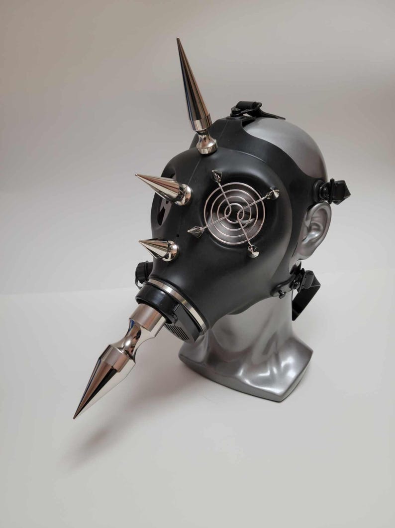 May include: A black gas mask with silver metal spikes and accents. The mask has a circular fan design on the front and is displayed on a gray mannequin head. The mask has adjustable straps.