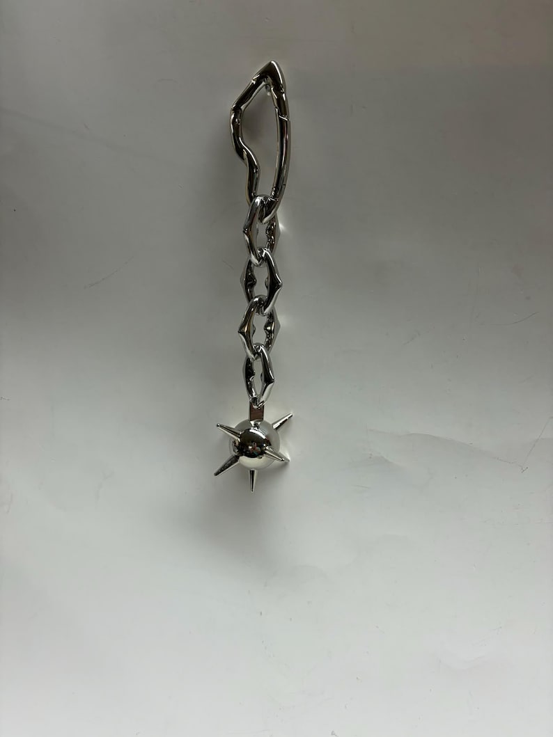 May include: A silver-toned metal earring featuring an irregular, elongated top link connected to a short chain of three hexagonal links, ending in a spherical charm with six protruding spikes extending from its surface in a star-like pattern.
