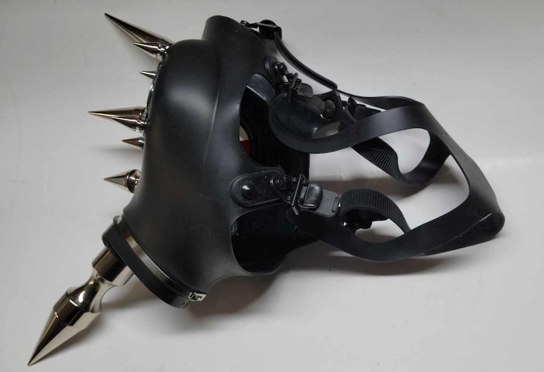 May include: Black gas mask with multiple silver metal spikes attached. The mask has adjustable black straps and a silver metal nozzle. The mask is made of a rubbery material and is designed to cover the face.