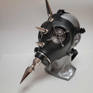 May include: A black gas mask with silver spikes and accents. The mask features a silver nose piece and a small fan-like detail on the front. The mask is displayed on a gray mannequin head.