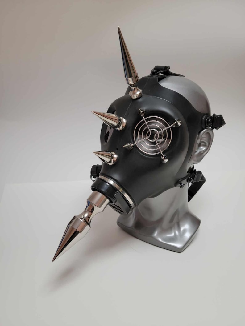 May include: A black gas mask with silver spikes and accents. The mask features a silver nose piece and a small fan-like detail on the front. The mask is displayed on a gray mannequin head.