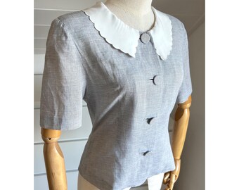 May include: A light gray short-sleeved blouse with a white scalloped collar and three matching buttons. The blouse is displayed on a mannequin with wooden arms and a white background.