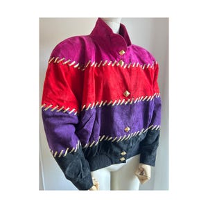 Peut inclure: Veste en daim colorée aux tons rose, rouge, violet et noir. La veste présente des coutures dorées et des boutons carrés dorés. Le col est montant et les poignets sont côtelés.