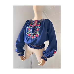 Könnte beinhalten: Eine marineblaue Bluse mit langen Ärmeln und floraler Stickerei. Die Stickerei zeigt rote, gelbe und grüne Blumen. Die Bluse hat einen gerüschten Saum und Manschetten sowie einen Kordelzug am Ausschnitt mit roten Quasten.