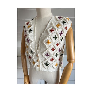 Può includere: Gilet senza maniche in maglia color crema con scollo a V e bottoni in legno. Il gilet presenta un motivo a rombi con disegni a croce ricamati colorati in rosso, verde, giallo e viola. Capo di ispirazione vintage.