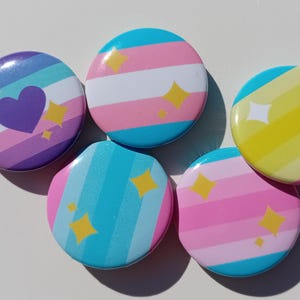 May include: Five colourful pin badges with various designs. The badges feature stripes, hearts, and stars in blue, pink, yellow, and purple. The badges are round and have a glossy finish, with a variety of LGBTQ+ pride flag designs.