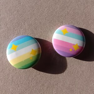 Peut inclure: Deux badges ronds. L'un présente un motif à rayures arc-en-ciel avec des étoiles jaunes. L'autre a un motif à rayures roses, violettes et blanches avec des étoiles jaunes. Les badges sont sur une surface marron clair.