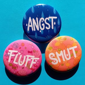 Pode incluir: Três botões redondos com designs e textos diferentes. O primeiro botão é azul com gotas de chuva brancas e o texto "ANGST". O segundo botão é rosa com doces brancos e o texto "FLUFF". O terceiro botão é laranja com frutas cítricas brancas e o texto "SMUT".