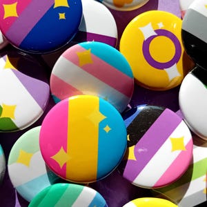 Może przedstawiać: Kolekcja kolorowych przypinek z różnymi flagami i symbolami dumy. Przypinki mają różne kolory, paski i wzory, w tym flagę transpłciową, flagę biseksualną i flagę panseksualną.