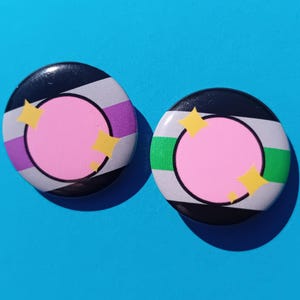 Op de afbeelding: Twee ronde buttons met een roze centrum, zwart-witte strepen en gele steraccenten. Eén button heeft paarse accenten, de andere groen. De buttons liggen op een helderblauwe achtergrond.