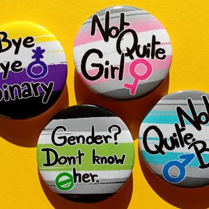 Può includere: Quattro bottoni rotondi con diversi design e testi. Il primo bottone ha uno sfondo a strisce giallo, viola e nero con il testo "Bye Bye Binary". Il secondo bottone ha uno sfondo a strisce grigio, rosa e bianco con il testo "Not Quite Girl". Il terzo bottone ha uno sfondo a strisce grigio, verde e nero con il testo "Gender? Don't know Other.". Il quarto bottone ha uno sfondo a strisce grigio, blu e bianco con il testo "Quite Not Boy".