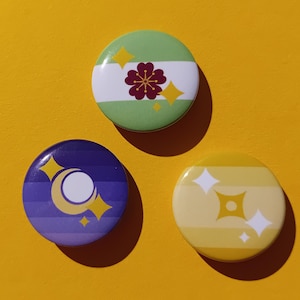 Può includere: Tre spille rotonde con disegni colorati. Una è verde con un fiore e strisce bianche, un'altra è viola con una luna e stelle, e l'ultima è gialla con esplosioni di stelle. Le spille sono su sfondo giallo.