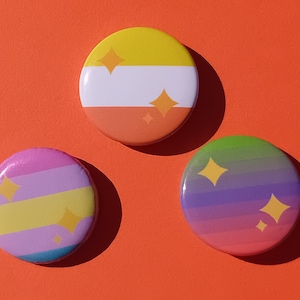 May include: Three colorful round buttons with glittery star accents on an orange background. One button has yellow, white, and orange stripes. Another has pink, yellow, and blue stripes. The third button has a rainbow gradient.