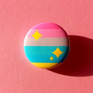 May include: A round button with a striped design in pink, gray, teal, and yellow. Two yellow starbursts are on the teal section, and one starburst is on the pink section. The button is on a pink background.