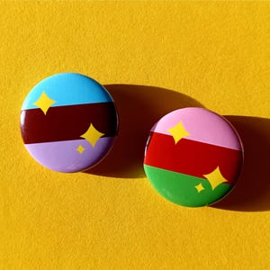 Puede incluir: Dos botones redondos y coloridos sobre un fondo amarillo. Un botón presenta secciones azules, granates y lavanda con acentos de estrellas amarillas. El otro tiene secciones rosas, rojas y verdes con acentos de estrellas amarillas.