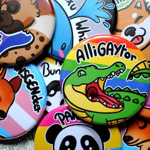 May include: A collection of colourful round buttons with various designs. The buttons feature illustrations of animals, including a deer, a fox, a panda, and an alligator. The buttons also include text, such as "Misgendered", "Bun", "Alligaytor", and "Whiskers".