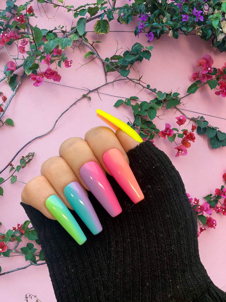 Op de afbeelding: Een hand met lange, coffin-vormige acrylnagels. De nagels zijn geschilderd in een kleurengradi&euml;nt, van groen naar blauw, paars, roze en oranje. De duimnagel is felgeel. De achtergrond is een roze muur met groene wijnranken en roze bloemen.