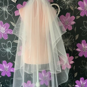 May include: A sheer, white bridal veil with two layers and a beaded edge. The veil is draped over a mannequin. The background is a floral wallpaper with purple and grey flower patterns.
