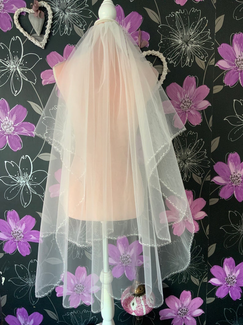 May include: A sheer, white bridal veil with two layers and a beaded edge. The veil is draped over a mannequin. The background is a floral wallpaper with purple and grey flower patterns.