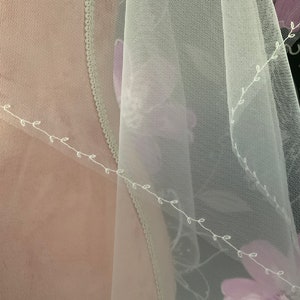 May include: A sheer, white veil with delicate floral embroidery and a scalloped edge. The veil features a soft, flowing fabric with a subtle floral pattern in shades of purple. The design includes a delicate vine-like embroidery.