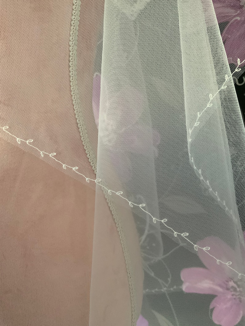 May include: A sheer, white veil with delicate floral embroidery and a scalloped edge. The veil features a soft, flowing fabric with a subtle floral pattern in shades of purple. The design includes a delicate vine-like embroidery.