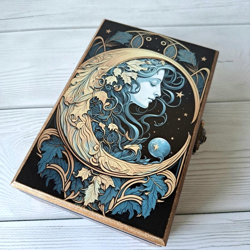 May include: A wooden box with a gold trim and a black background. The lid features a crescent moon with a woman's face inside, surrounded by blue and gold leaves. The moon has a small blue planet inside it.