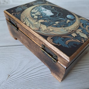 May include: A wooden box with a hinged lid. The lid is decorated with a dark blue and gold illustration of a woman's face surrounded by leaves and a crescent moon.
