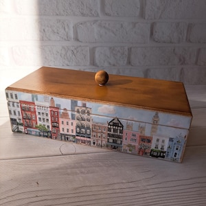 May include: A wooden box with a cityscape design. The box has a light brown wooden top and a cityscape design around the sides. The cityscape features buildings in various colors, including red, pink, and blue. A small, round wooden knob sits on top.