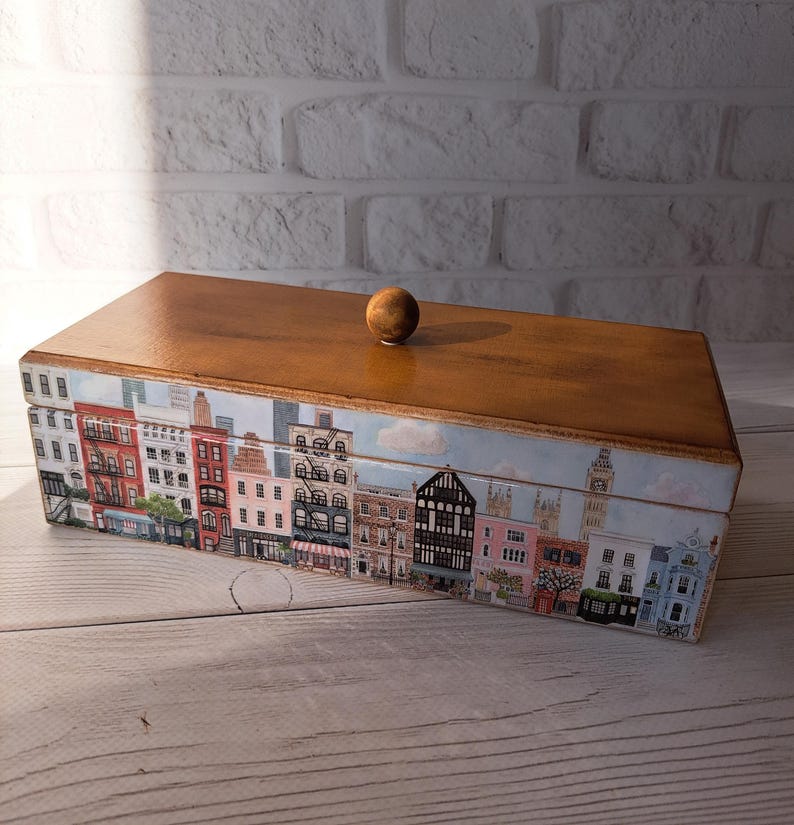 May include: A wooden box with a cityscape design. The box has a light brown wooden top and a cityscape design around the sides. The cityscape features buildings in various colors, including red, pink, and blue. A small, round wooden knob sits on top.