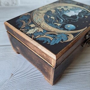 May include: A wooden box with a dark brown finish and a decorative lid featuring a moon, stars, and a woman's face. The lid is painted with blue, gold, and white details.