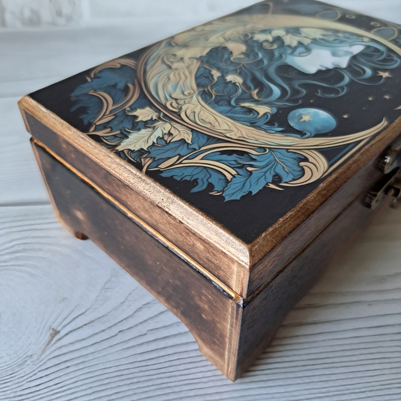 May include: A wooden box with a dark brown finish and a decorative lid featuring a moon, stars, and a woman's face. The lid is painted with blue, gold, and white details.