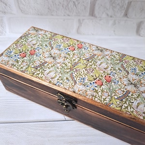 Può includere: Una scatola rettangolare in legno con coperchio a fantasia floreale. Il coperchio presenta uno sfondo color crema con un motivo ripetuto di gigli bianchi, fiori rossi e accenti blu. La scatola ha una finitura marrone scuro e una chiusura in metallo.