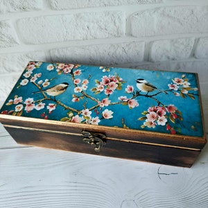 May include: A wooden box with a hinged lid and a metal latch. The lid is decorated with a blue background and a floral design featuring pink flowers and two birds.